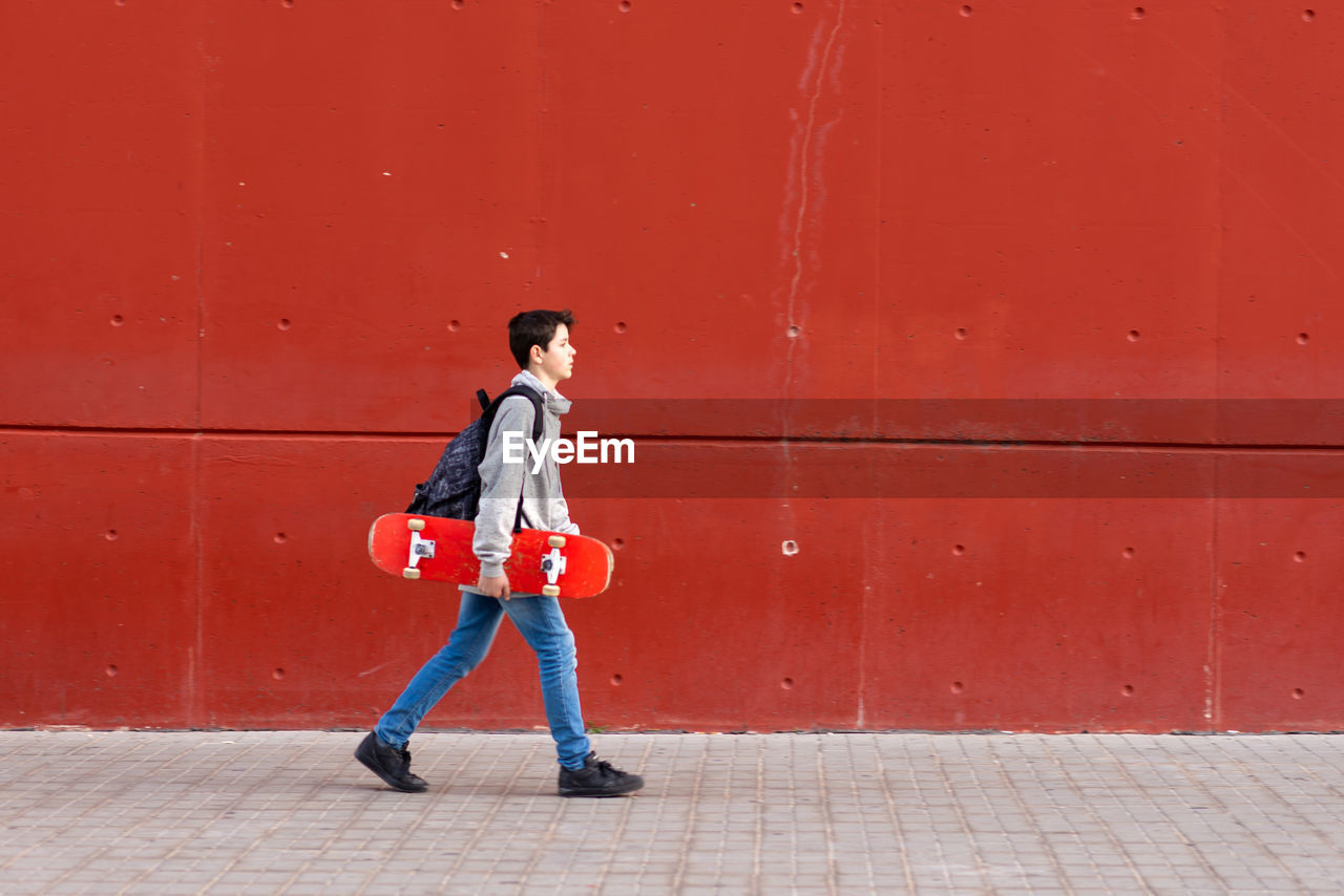 Young boy holding a red skateboard while walking against red wall person