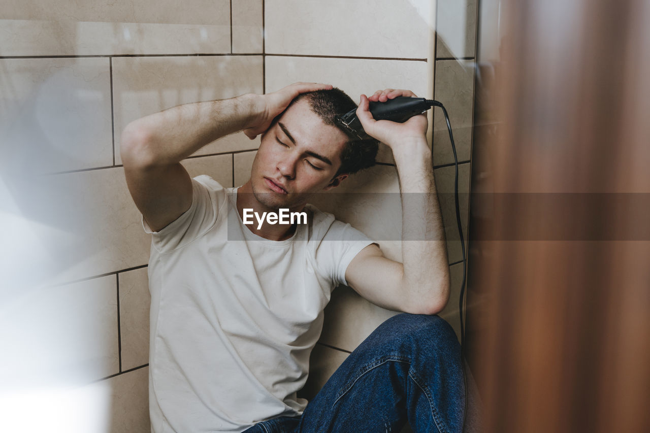 Depressed young man sitting with eyes closed shaving hair