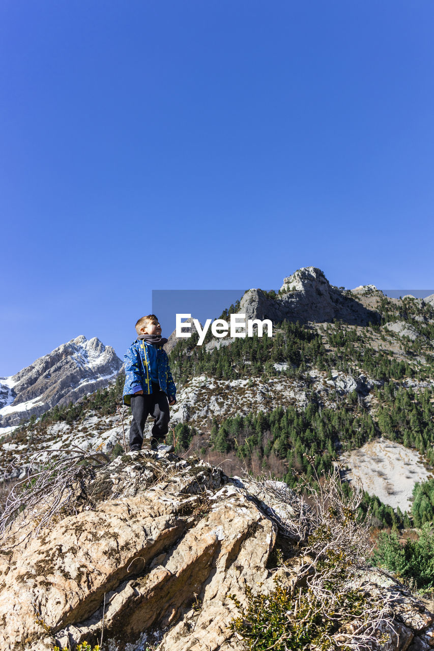 Boy standing on rock against clear blue sky