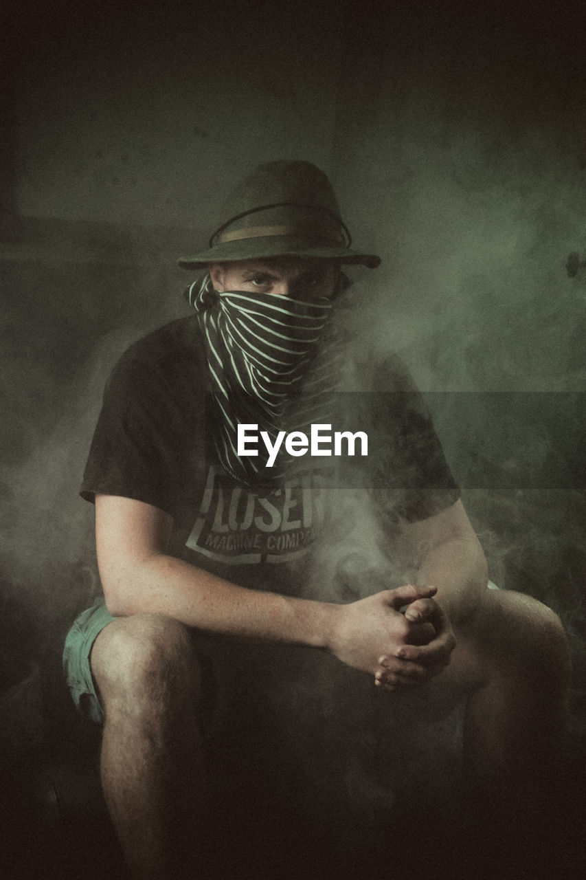 Portrait of man wearing hat and covering face sitting amidst smoke