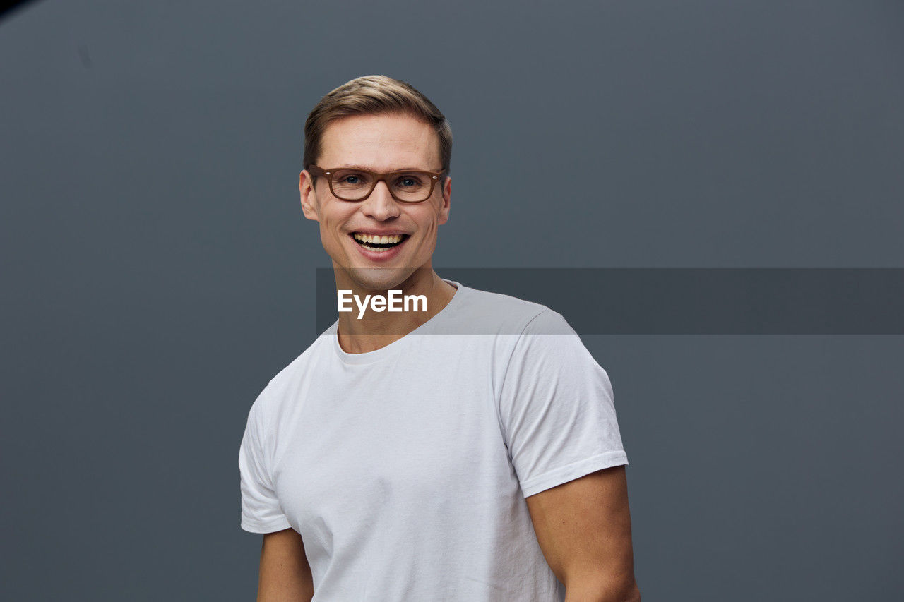 portrait, smiling, one person, eyeglasses, happiness, studio shot, looking at camera, glasses, casual clothing, adult, indoors, t-shirt, emotion, young adult, gray, front view, teeth, men, smile, standing, copy space, white, cheerful, waist up, person, gray background, beard, facial hair, positive emotion