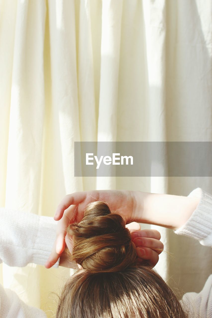 Cropped image of woman tying hair bun against curtain at home