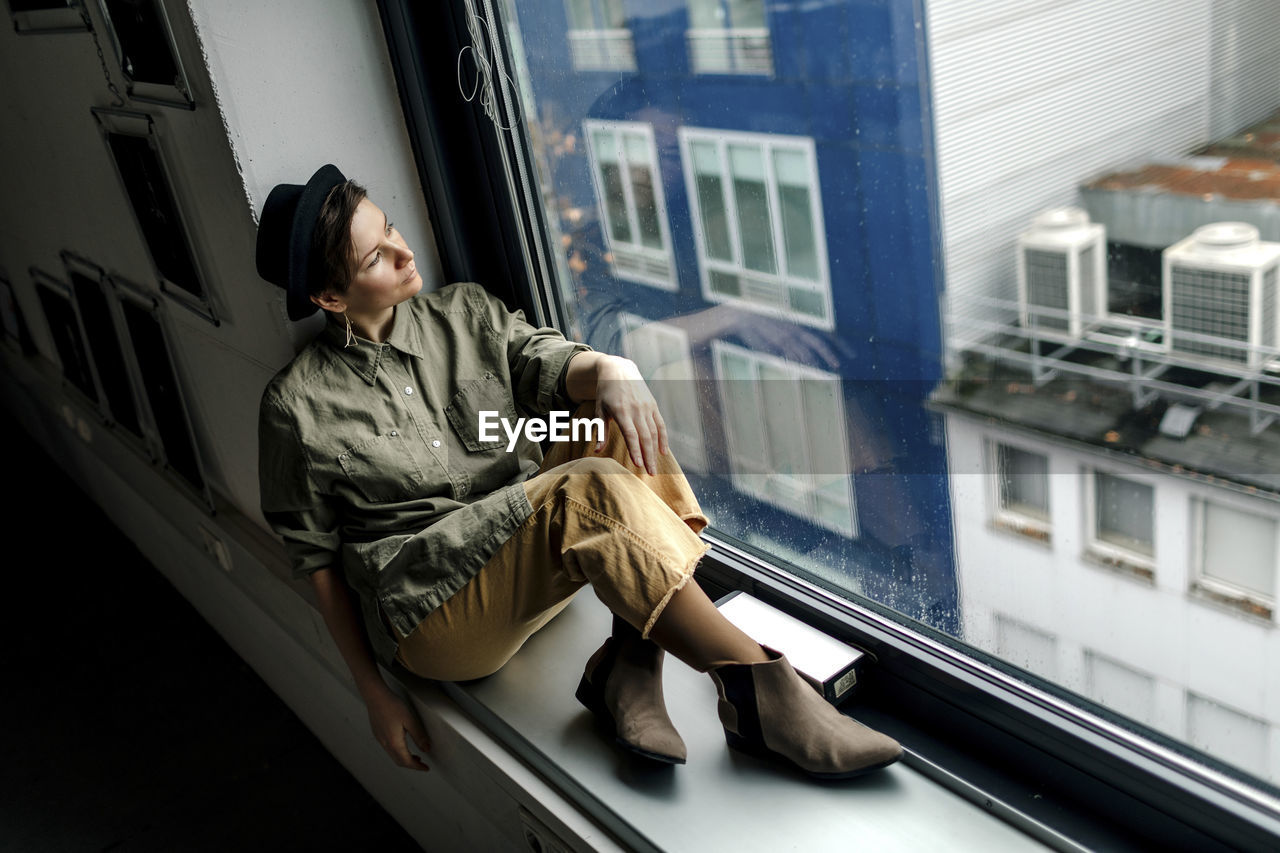 Fashionable woman looking through window while sitting on window sill at studio