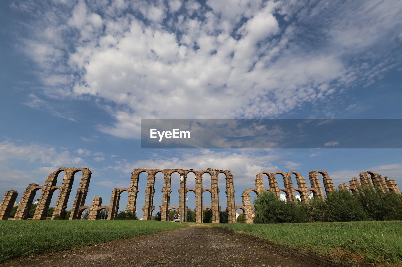 Wide angle of old ruin, roman aqueduct on field against blue sky with white clouds