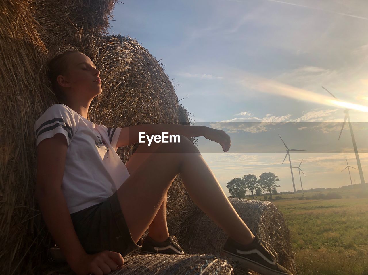 Girl sitting on hay bale against sky