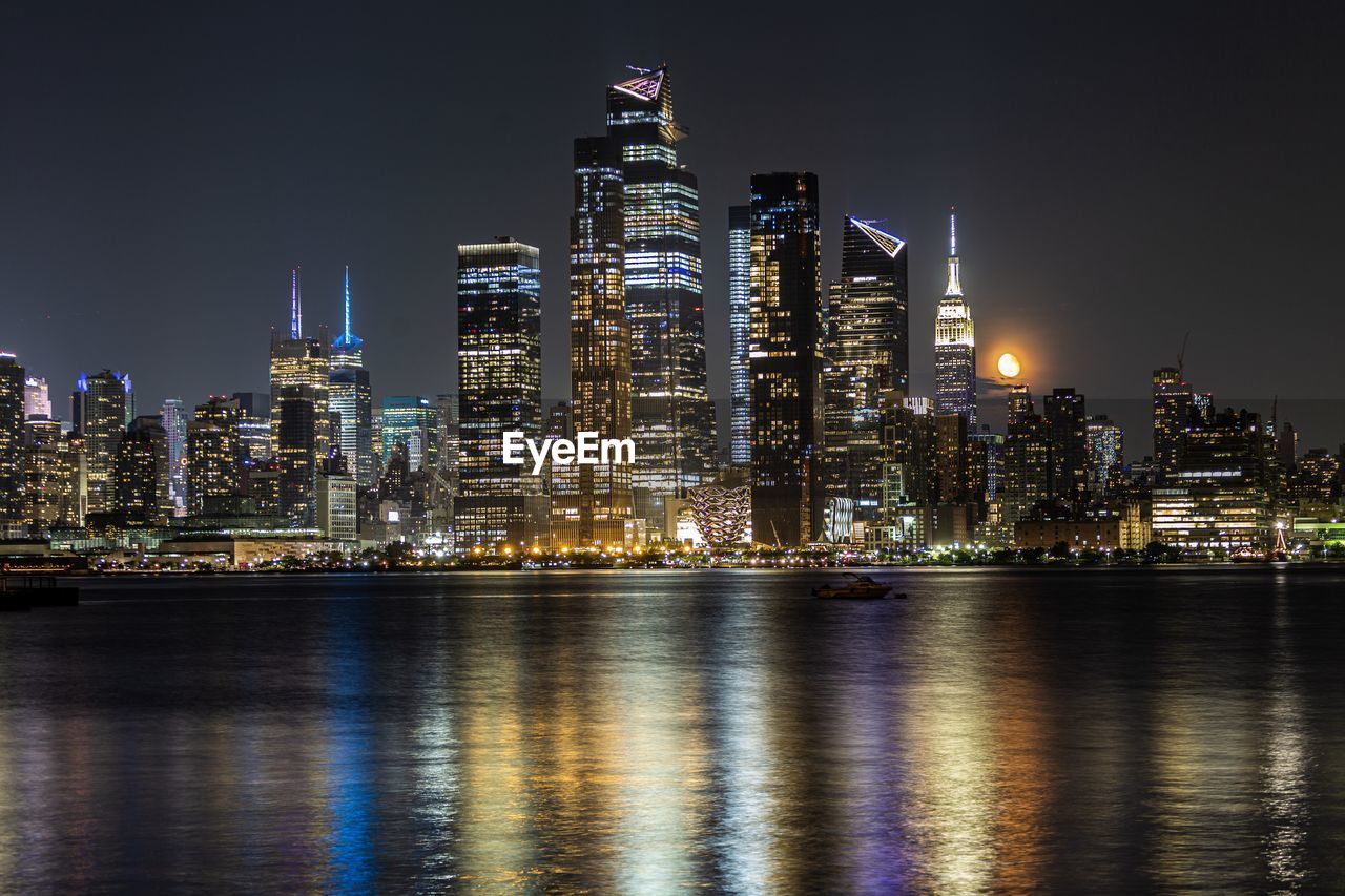 ILLUMINATED MODERN BUILDINGS BY RIVER AGAINST SKY AT NIGHT