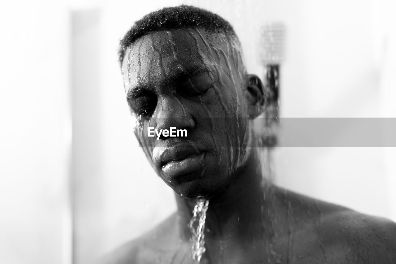 Black and white of emotionless young black guy taking shower in light bathroom with closed eyes and water on face