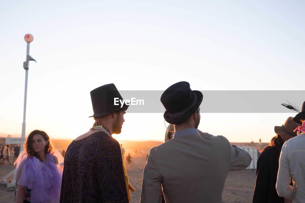 REAR VIEW OF PEOPLE STANDING AGAINST CLEAR SKY DURING SUNSET