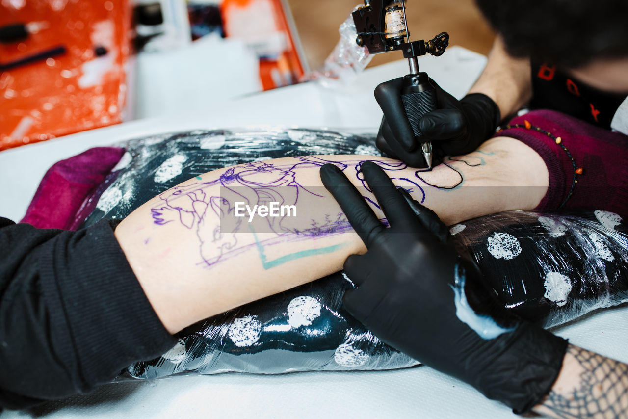 From above anonymous cropped tattooist in latex gloves using tattoo machine to make sketch on leg of crop client in modern salon  person