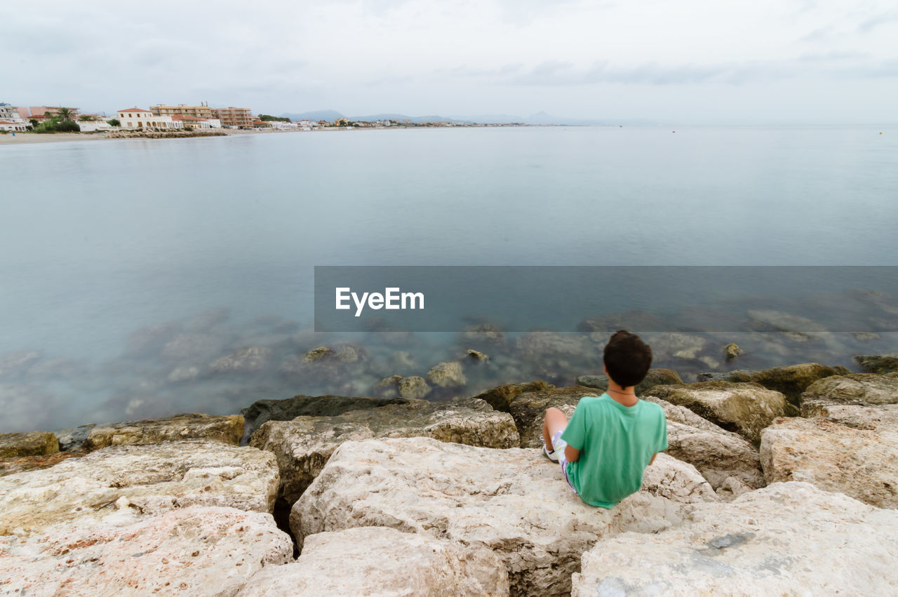 Rear view of boy looking away while sitting on rock by sea