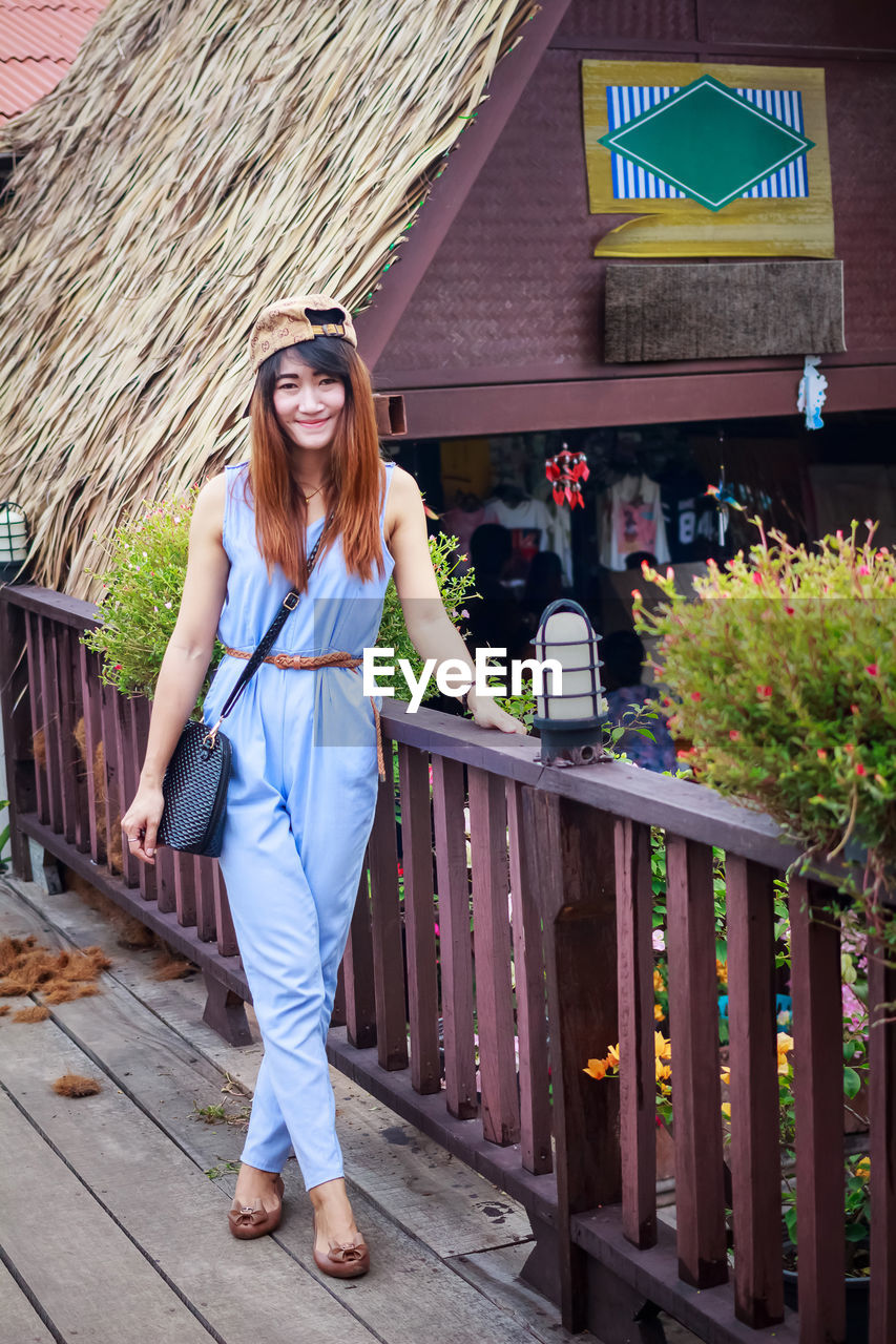 PORTRAIT OF SMILING YOUNG WOMAN STANDING AGAINST BUILT STRUCTURE