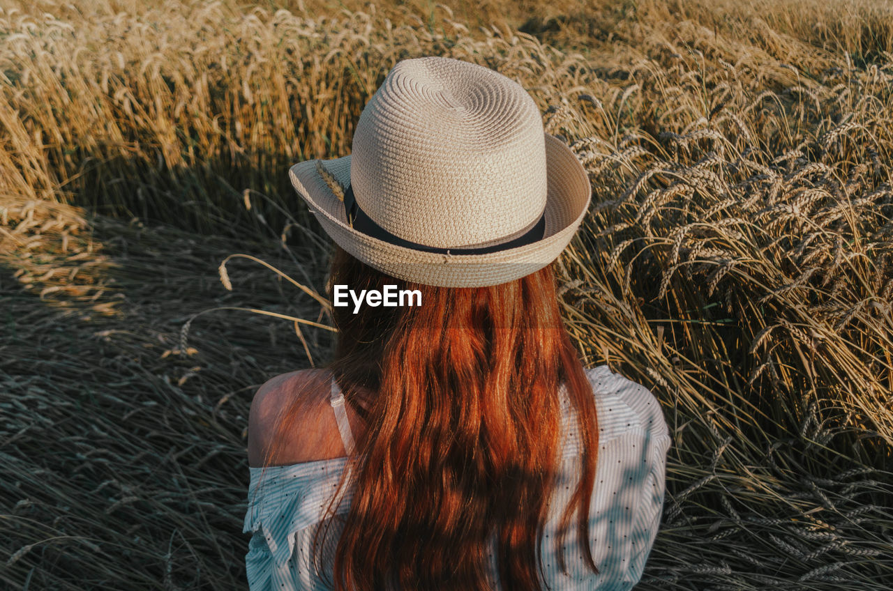 Rear view of woman wearing hat sitting amidst wheat crops at sunset