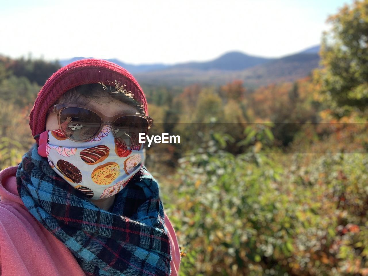 Portrait of young woman wearing sunglasses, face mask and scarf in front of fall landscape 