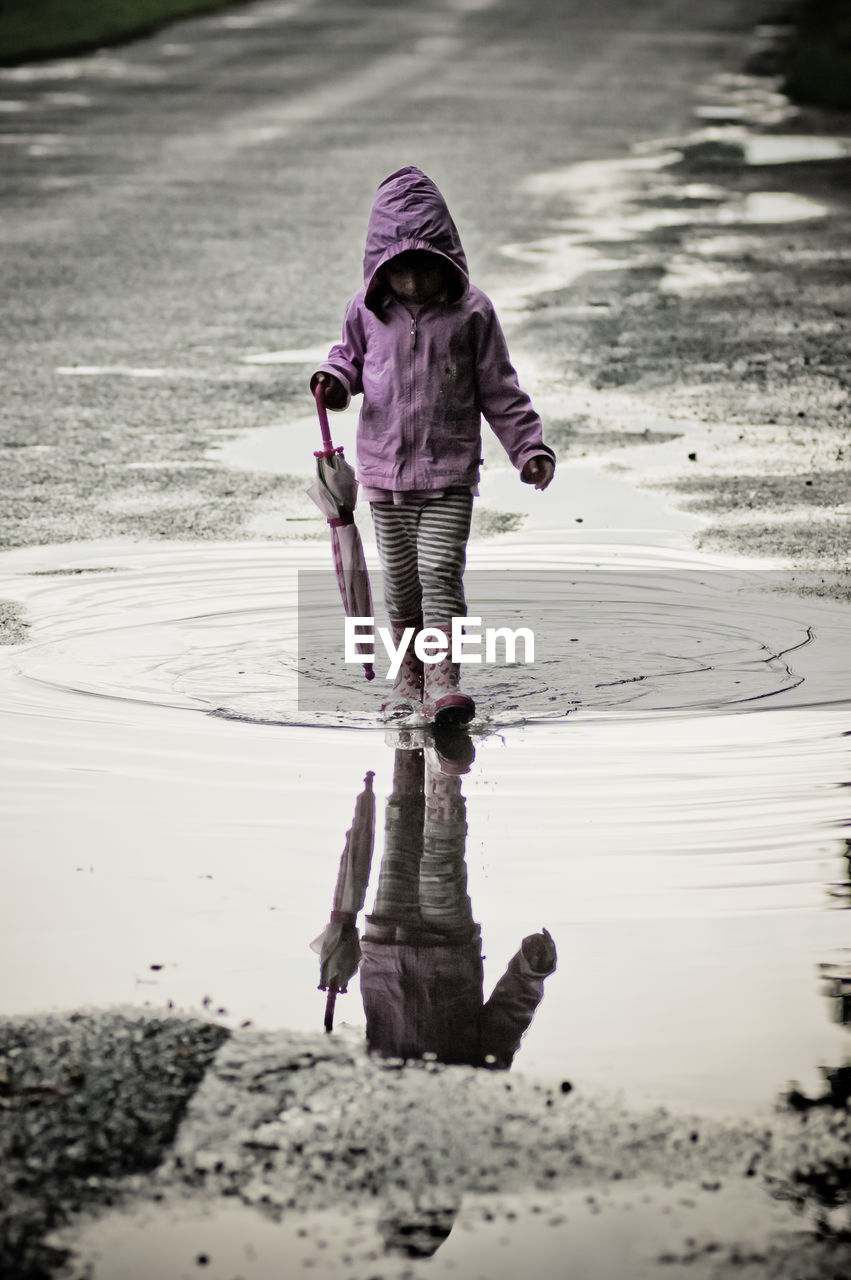 Full length of girl wearing raincoat and rubber boots while walking in puddle on road