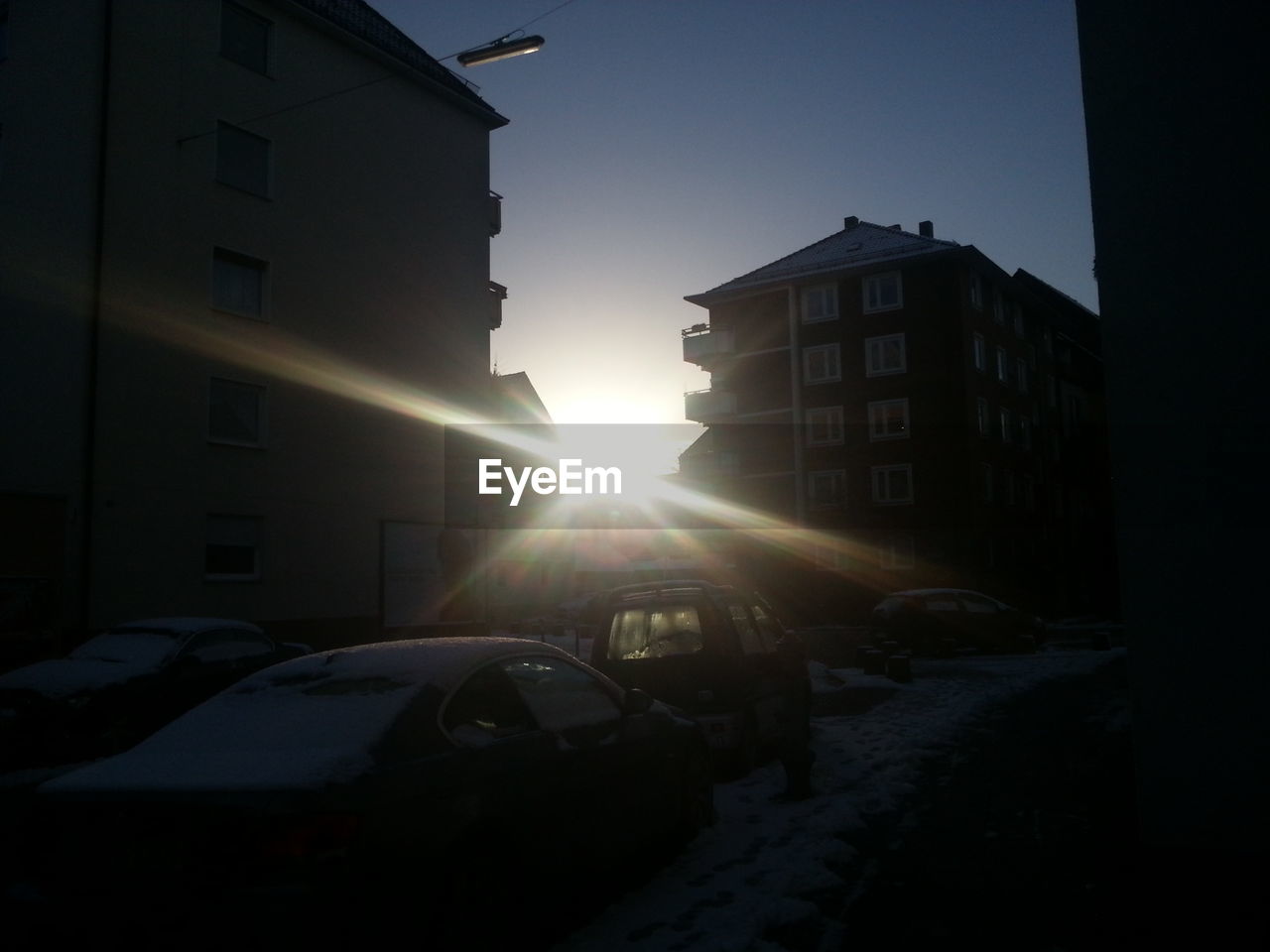 Cars on snow covered street in front of buildings during sunset