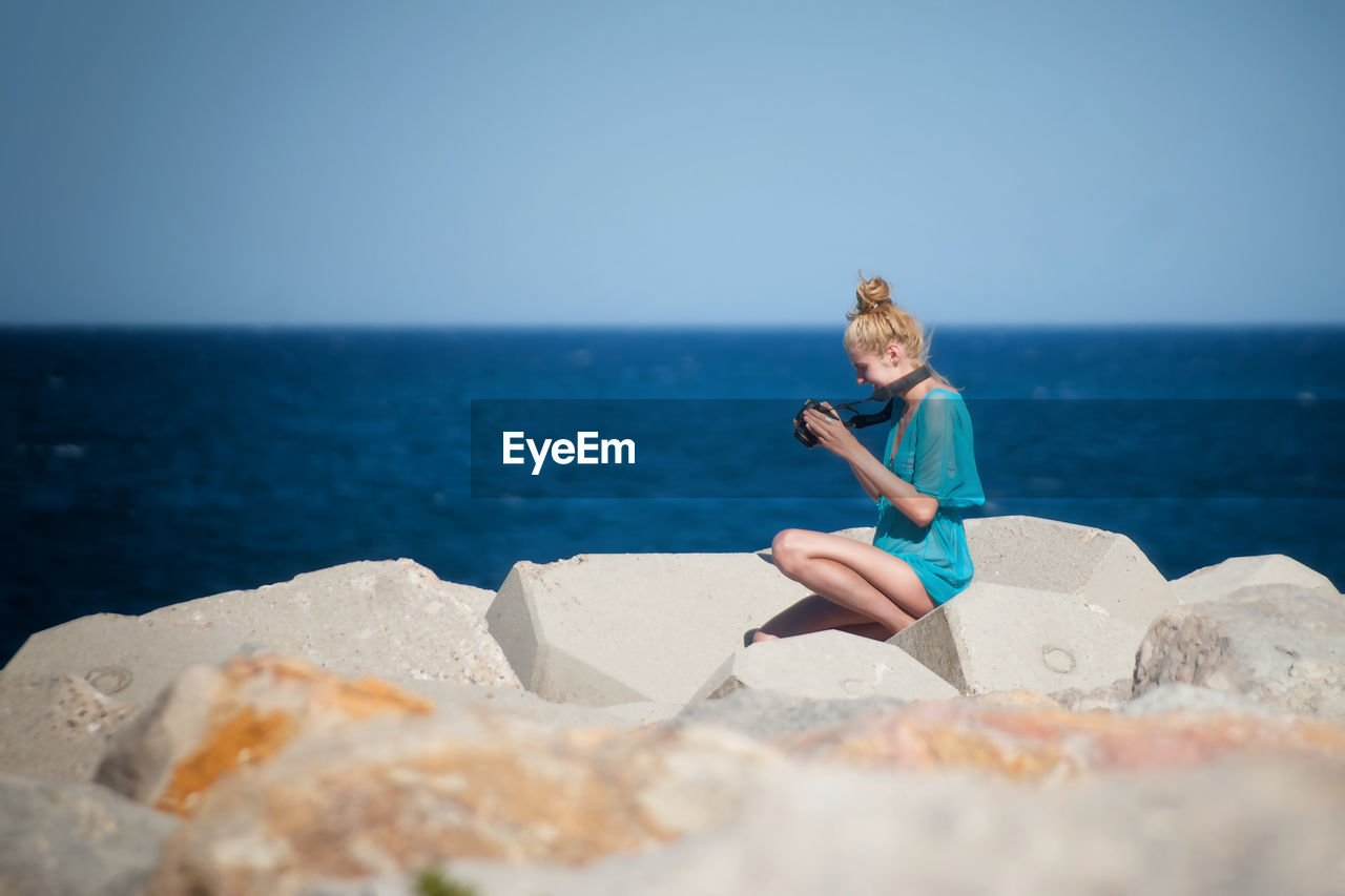 Side view of young woman looking at camera while sitting on rocks by sea