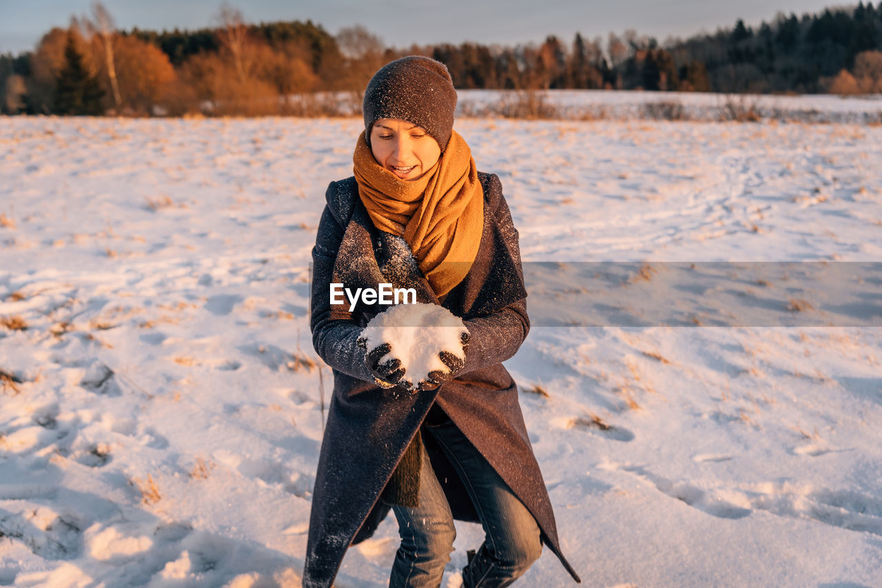 A woman in winter clothes collects a lump of snow in a snow-covered field