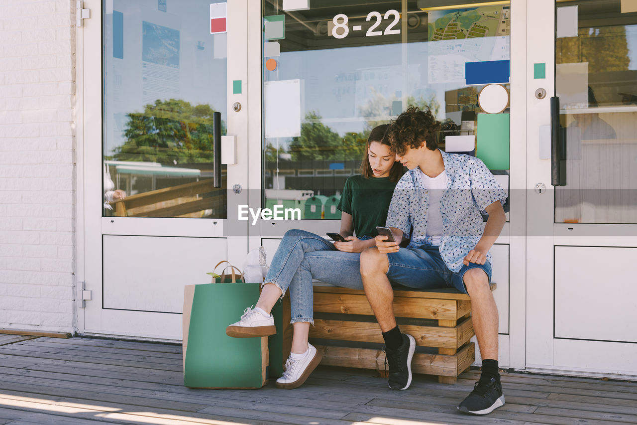 Siblings using mobile phone while sitting on bench with bags outside information booth