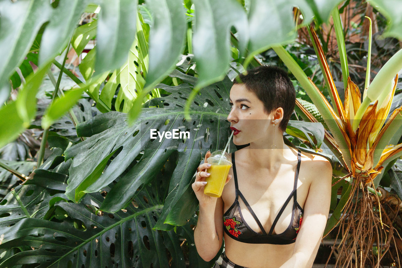 Young woman with short hair drinking juice while sitting against plants