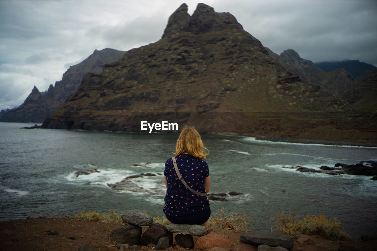 Rear view of woman sitting on rock by sea against mountains