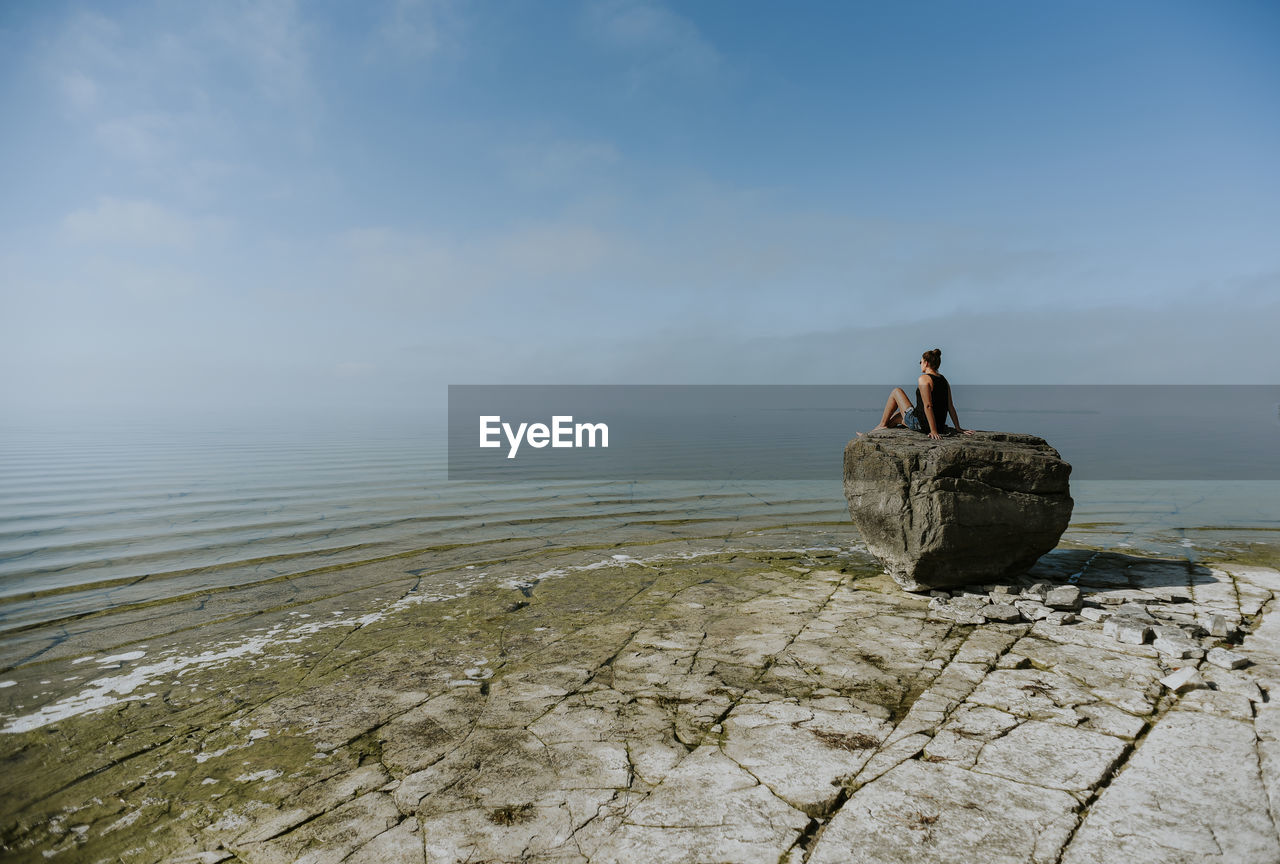 Woman looking at sea while sitting on rock against blue sky