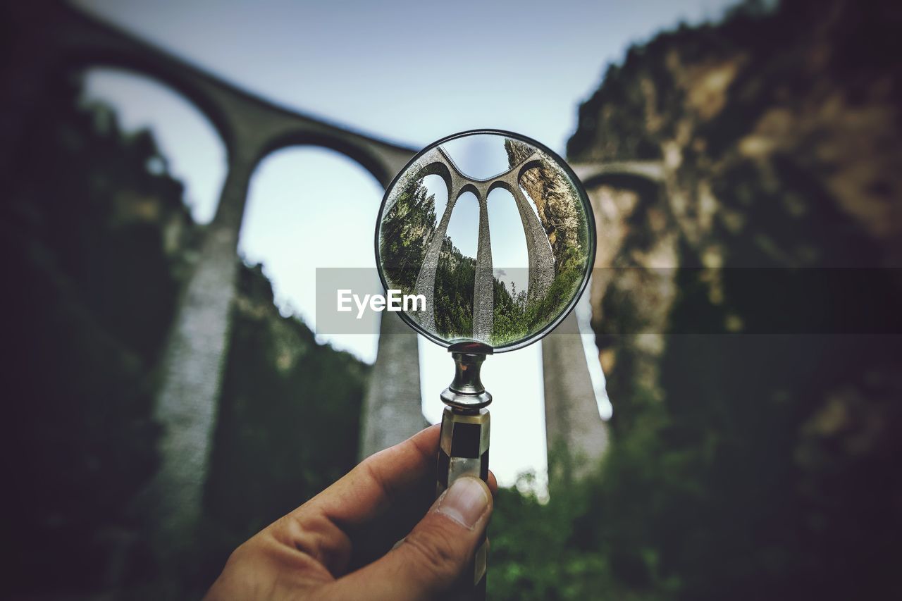 Cropped hand holding magnifying glass against viaduct bridge