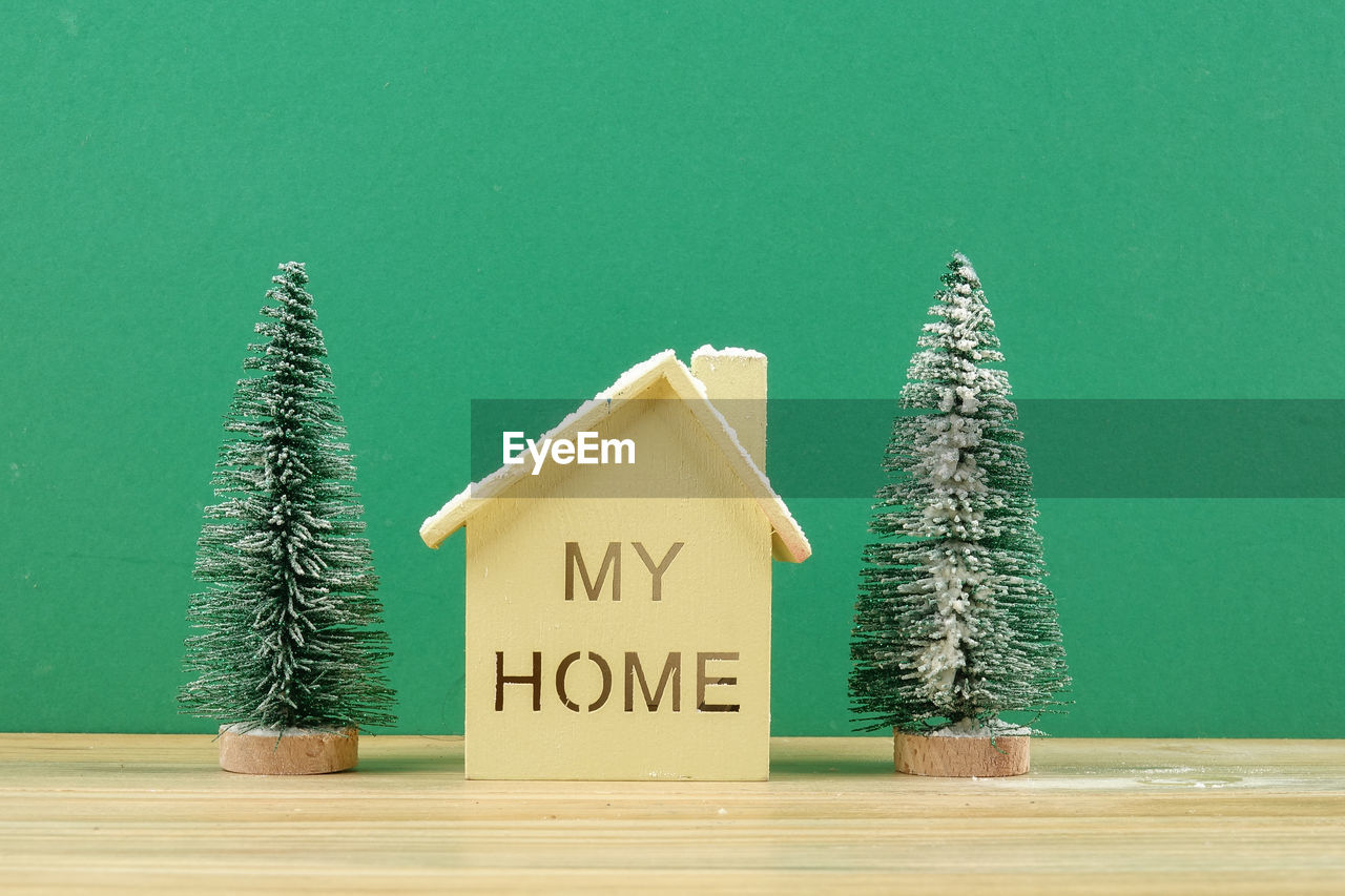 Close-up of model home with text artificial christmas trees on table against green background