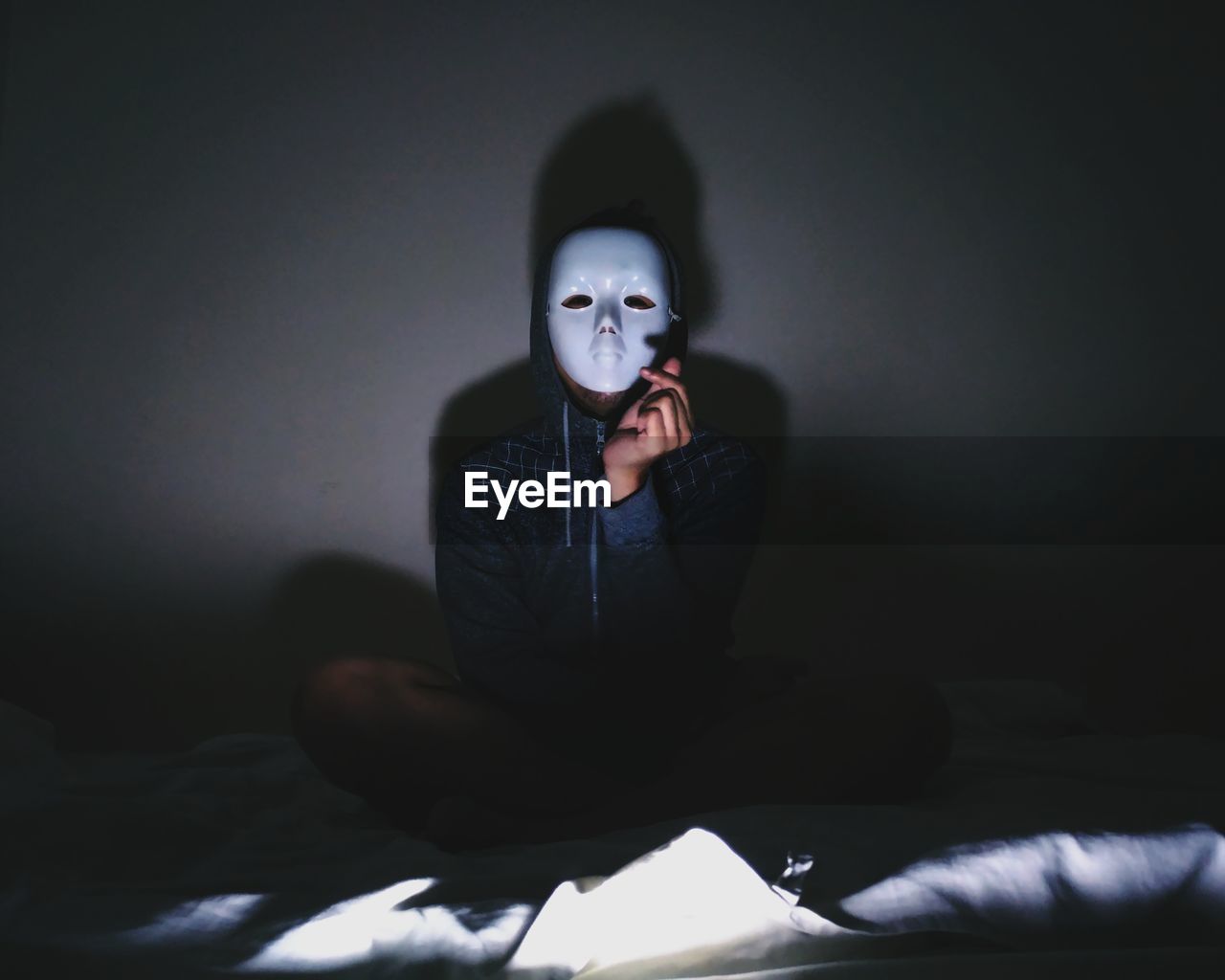 Portrait of young man wearing mask while sitting on bed in darkroom