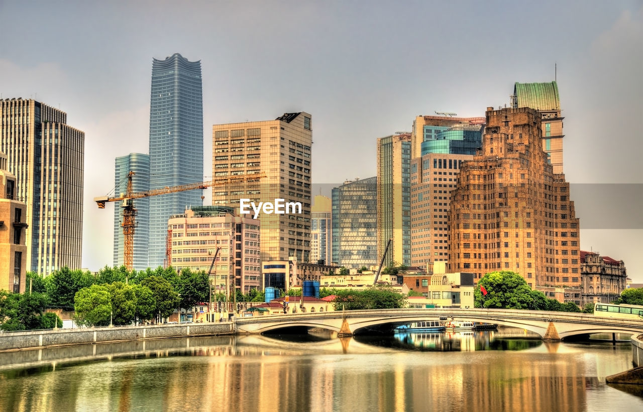 BRIDGE OVER RIVER AMIDST BUILDINGS AGAINST SKY IN CITY