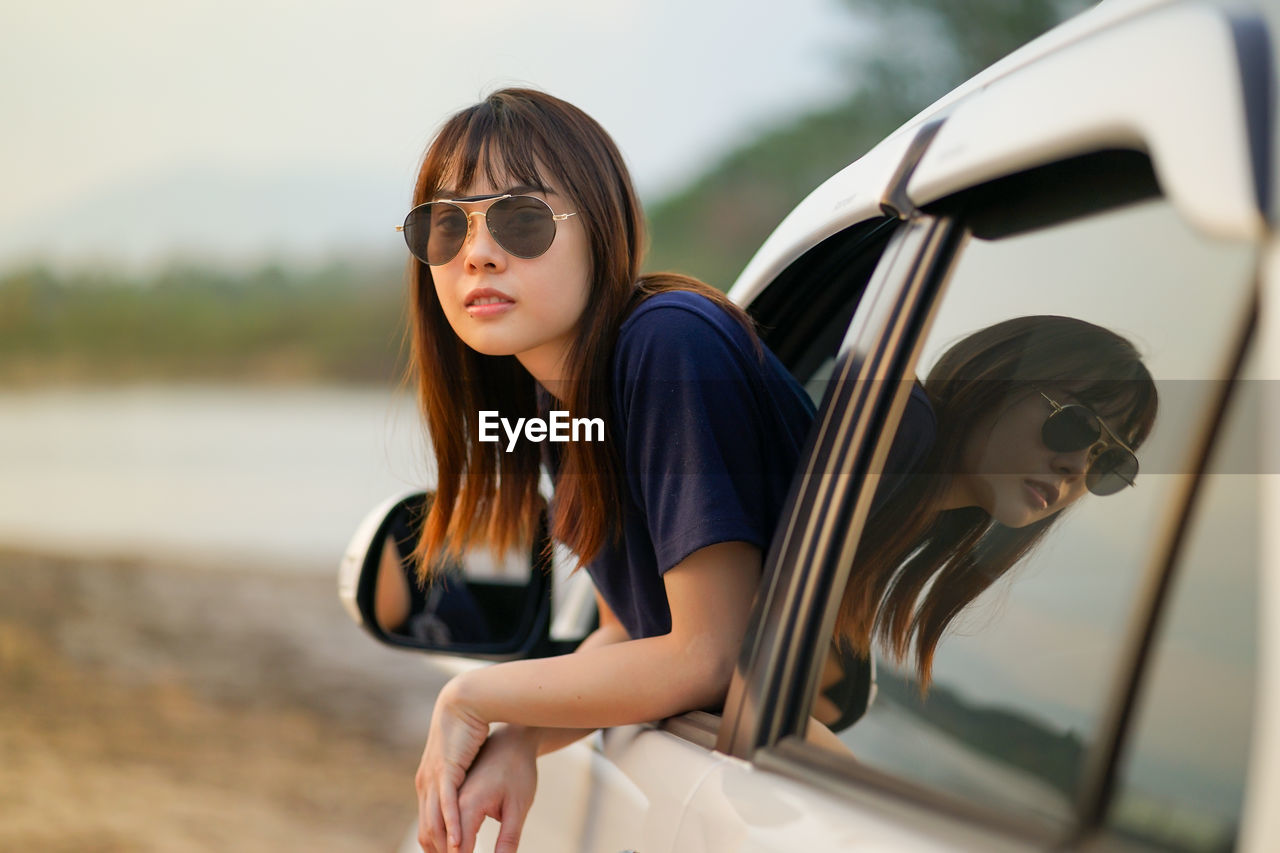 Portrait of young woman wearing sunglasses peeking through car window