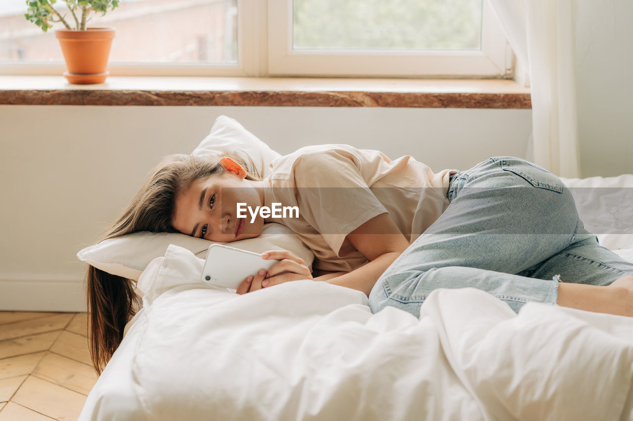 Sad young woman looking at her smartphone in the day while lying on the bed, internet addiction