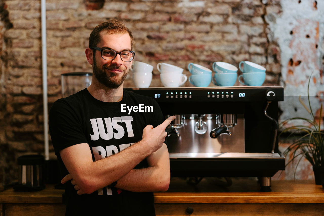Smiling male barista using portafilter and preparing coffee in modern coffeemaker while standing at counter in cafe and looking at camera