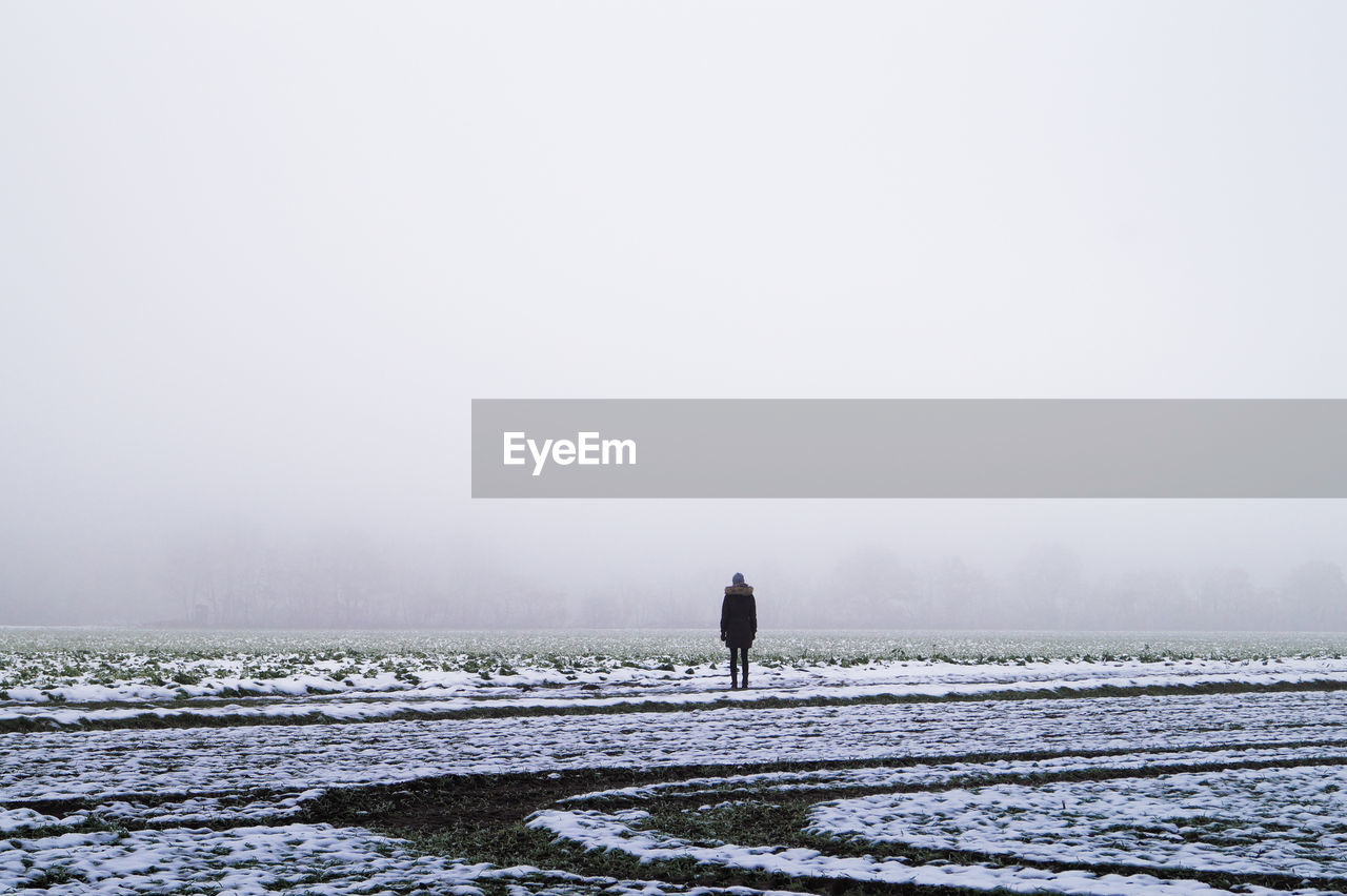 Mid distant rear view of person standing on snow covered field against sky during winter