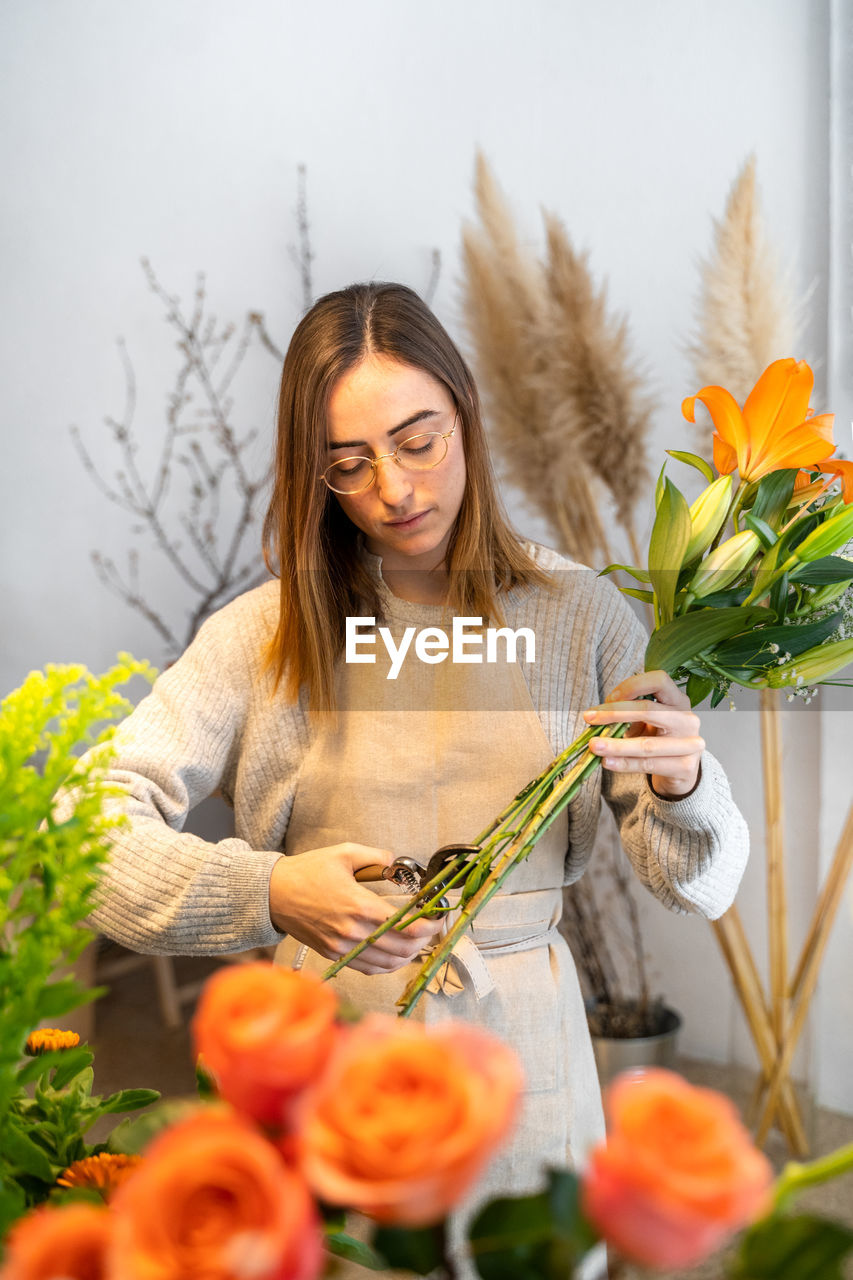 Focused young ethnic female florist in casual clothes and apron cutting stems of fresh bright flowers with pruning snips in floral shop