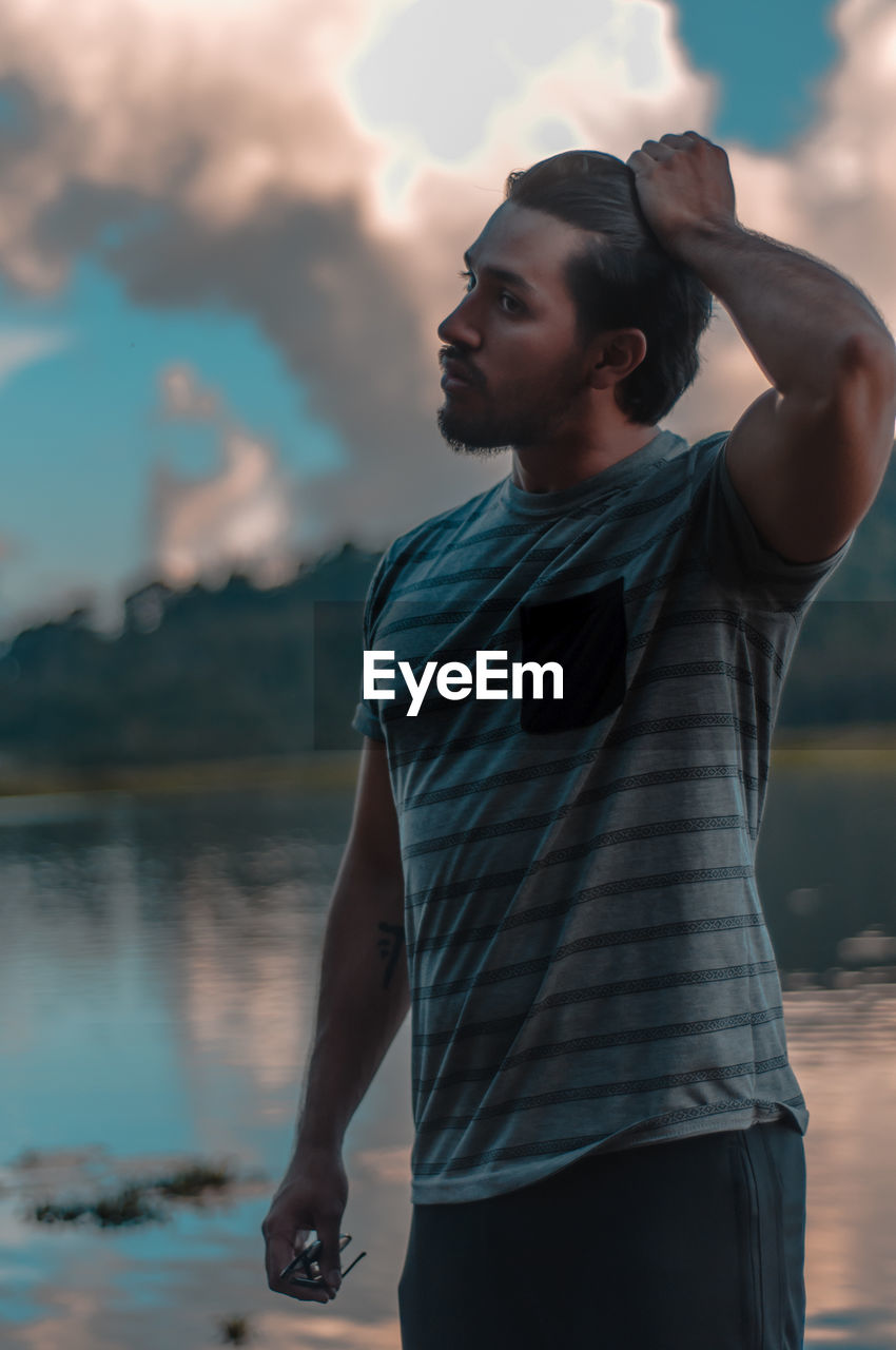 Young man looking away while standing on lake against sky