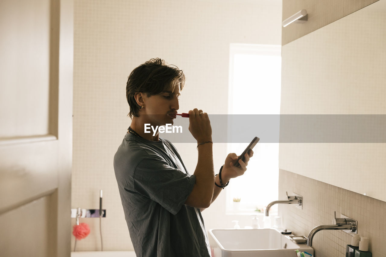 Side view of young man using smart phone while brushing teeth in bathroom at home