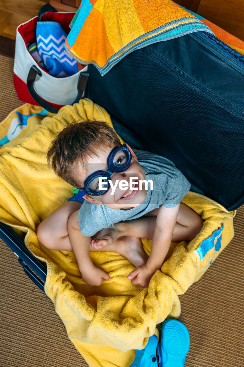 High angle portrait boy wearing swimming goggles while sitting in suitcase at home