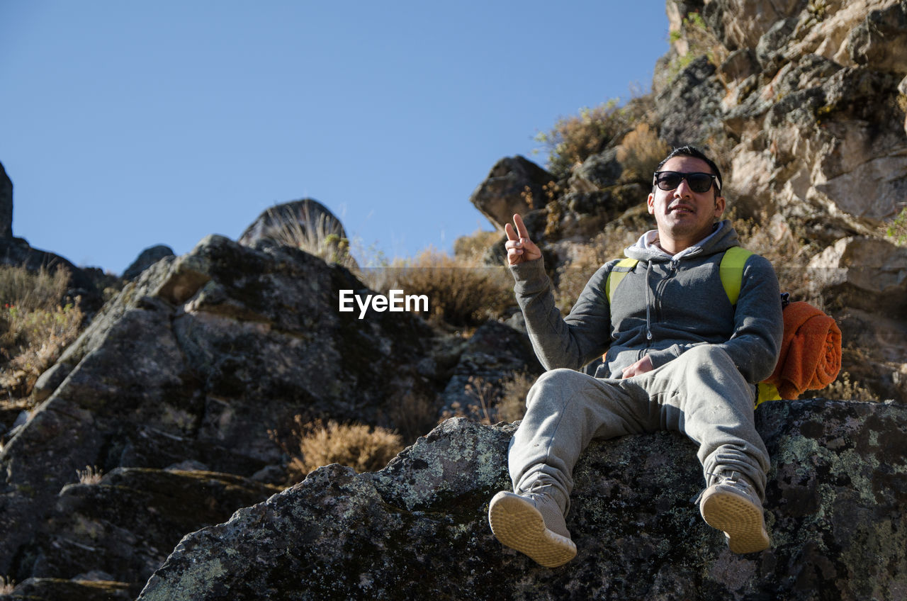 Low angle portrait of man gesturing while sitting on rock against mountains