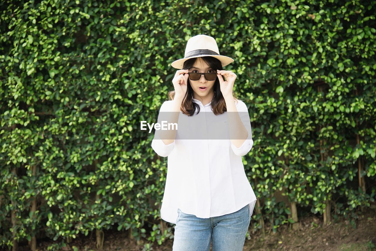 Portrait of young woman wearing hat and sunglasses while standing against plants