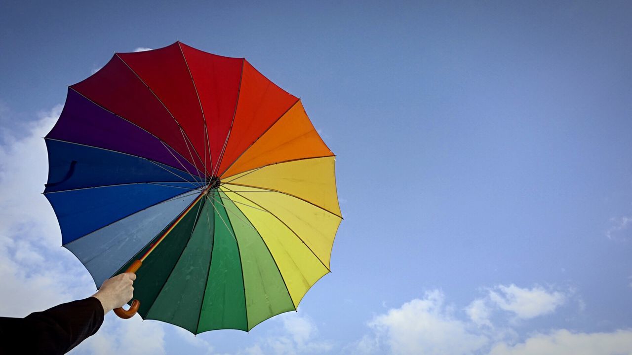 Cropped hand holding colorful umbrella against sky