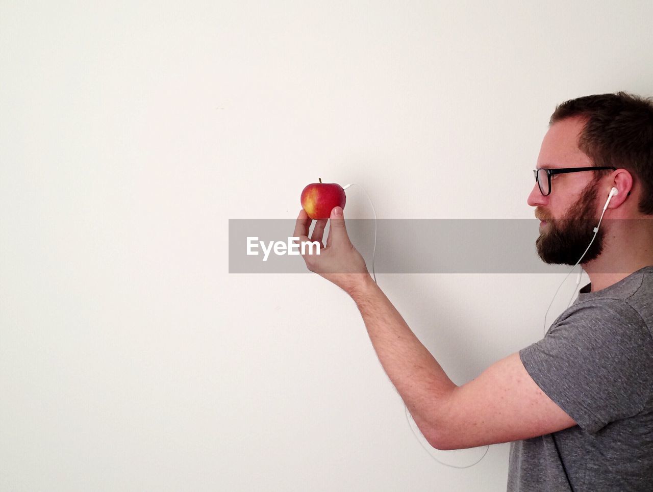 Side view of man holding apple with headphones against white wall