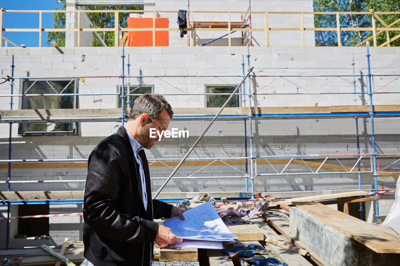 Side view of architect reading blueprint by incomplete building at construction site