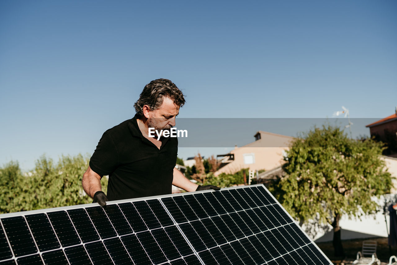 Mature technician man holding solar panels on house roof for self consumption energy. renewable