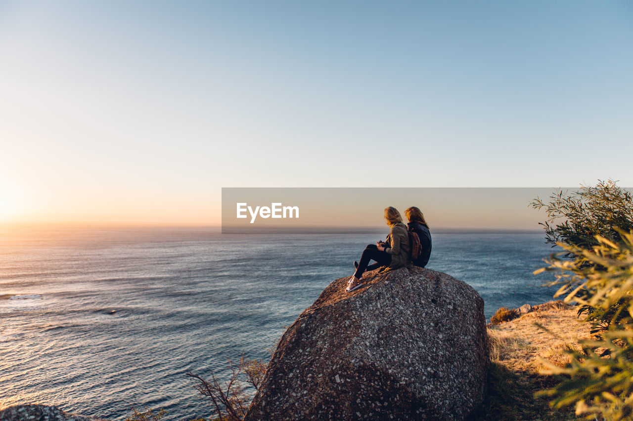 Side view of female friends sitting at beach against clear sky during sunset