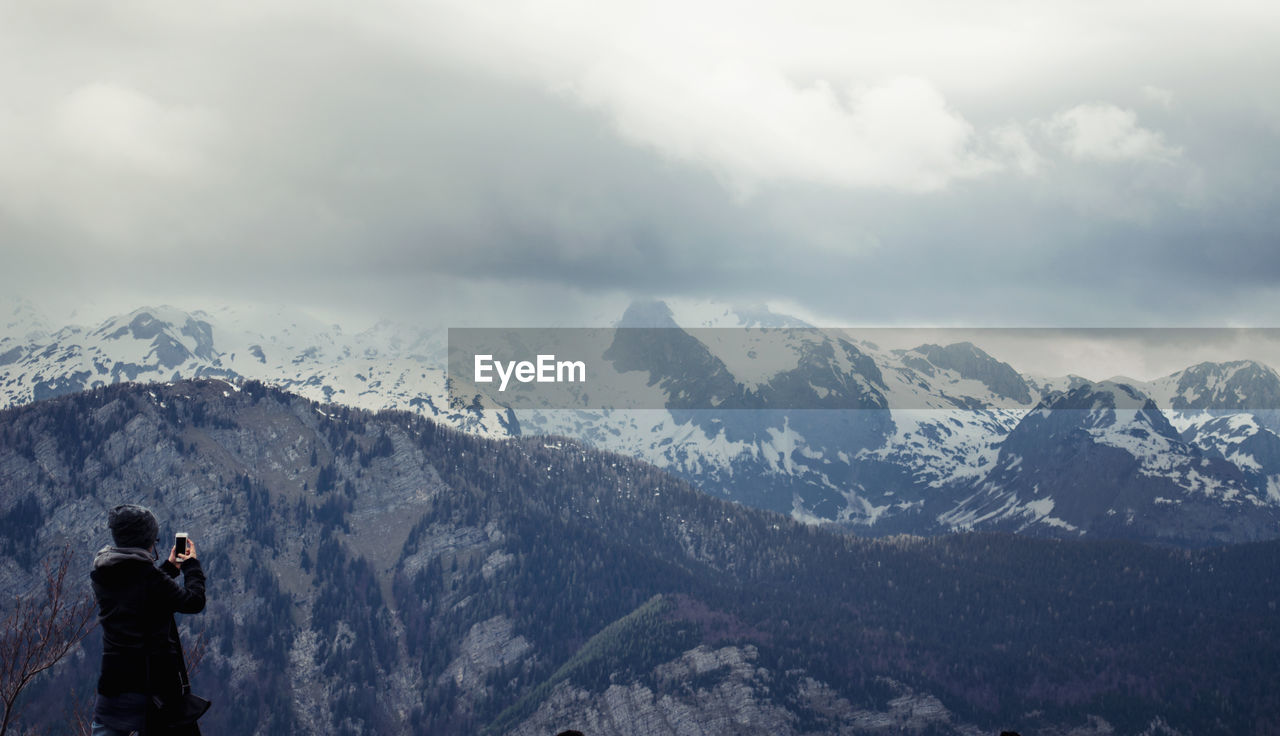 Rear view of photographing snow covered mountains against cloudy sky