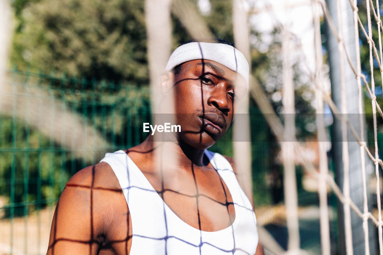 Serious african american male athlete in white activewear standing near net on playground in summer and looking at camera