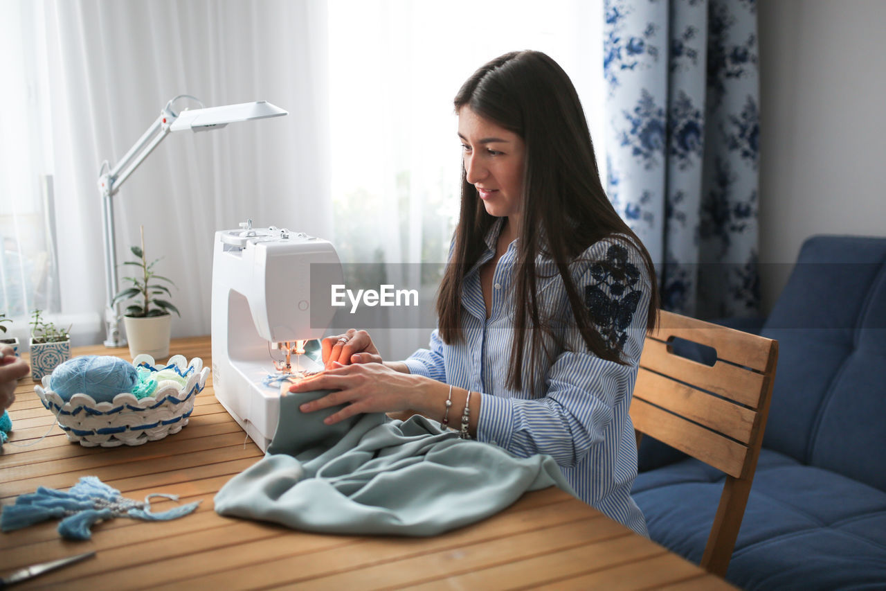 Beautiful woman with long dark hair sews a blue cloth on a sewing machine. 