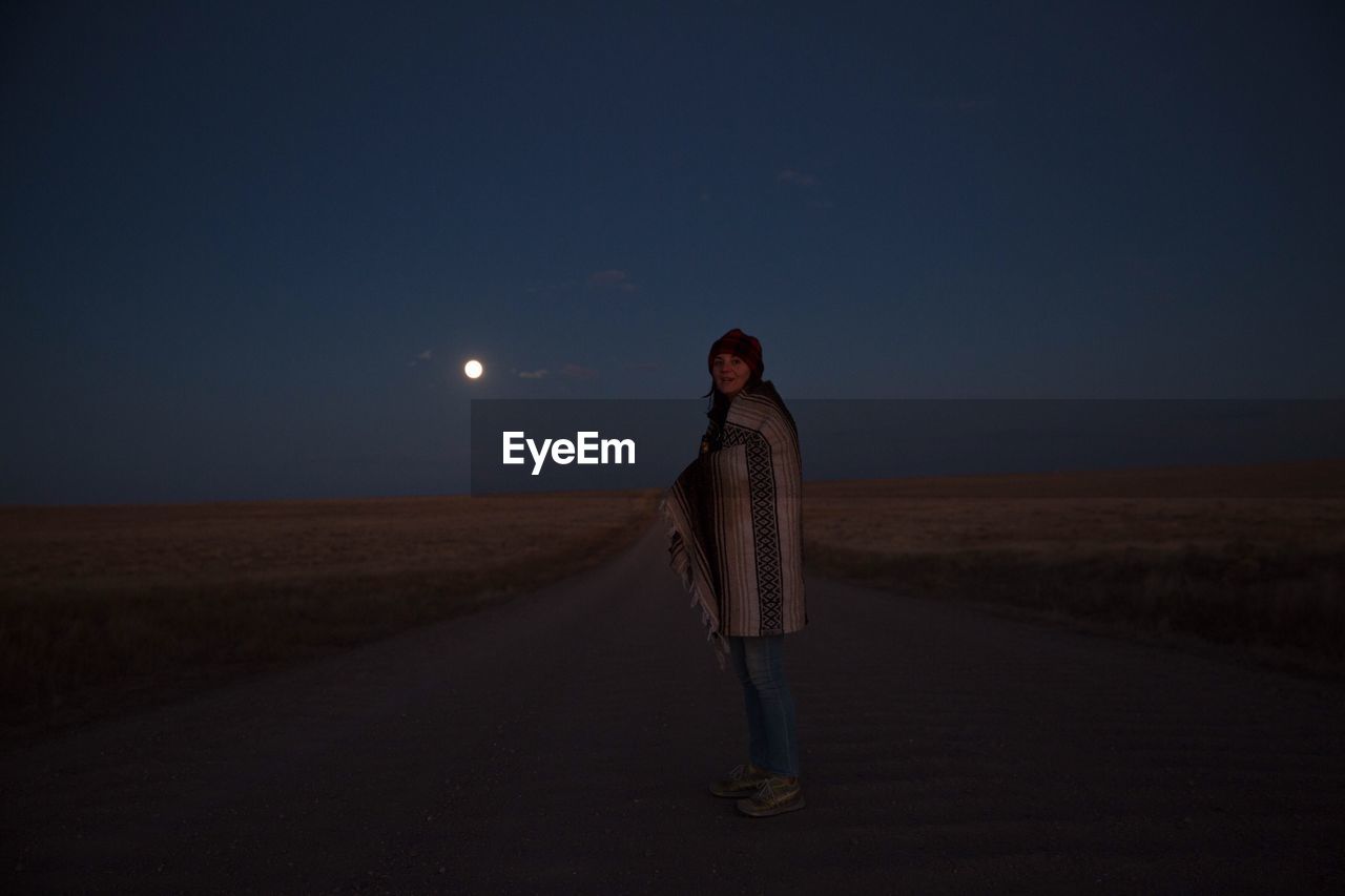 Woman wrapped in blanket standing on street amidst field against sky at dusk