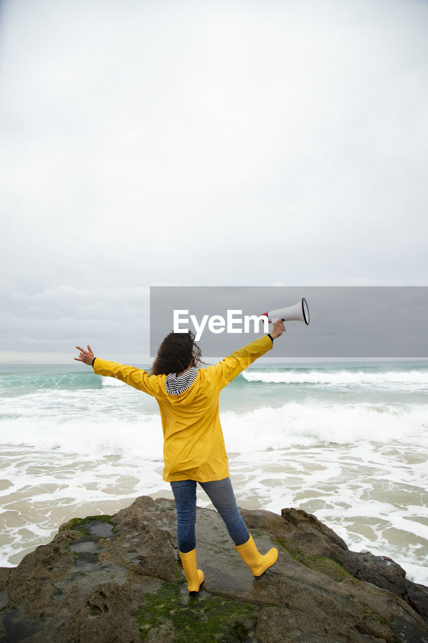 Carefree young woman holding megaphone while standing against sea