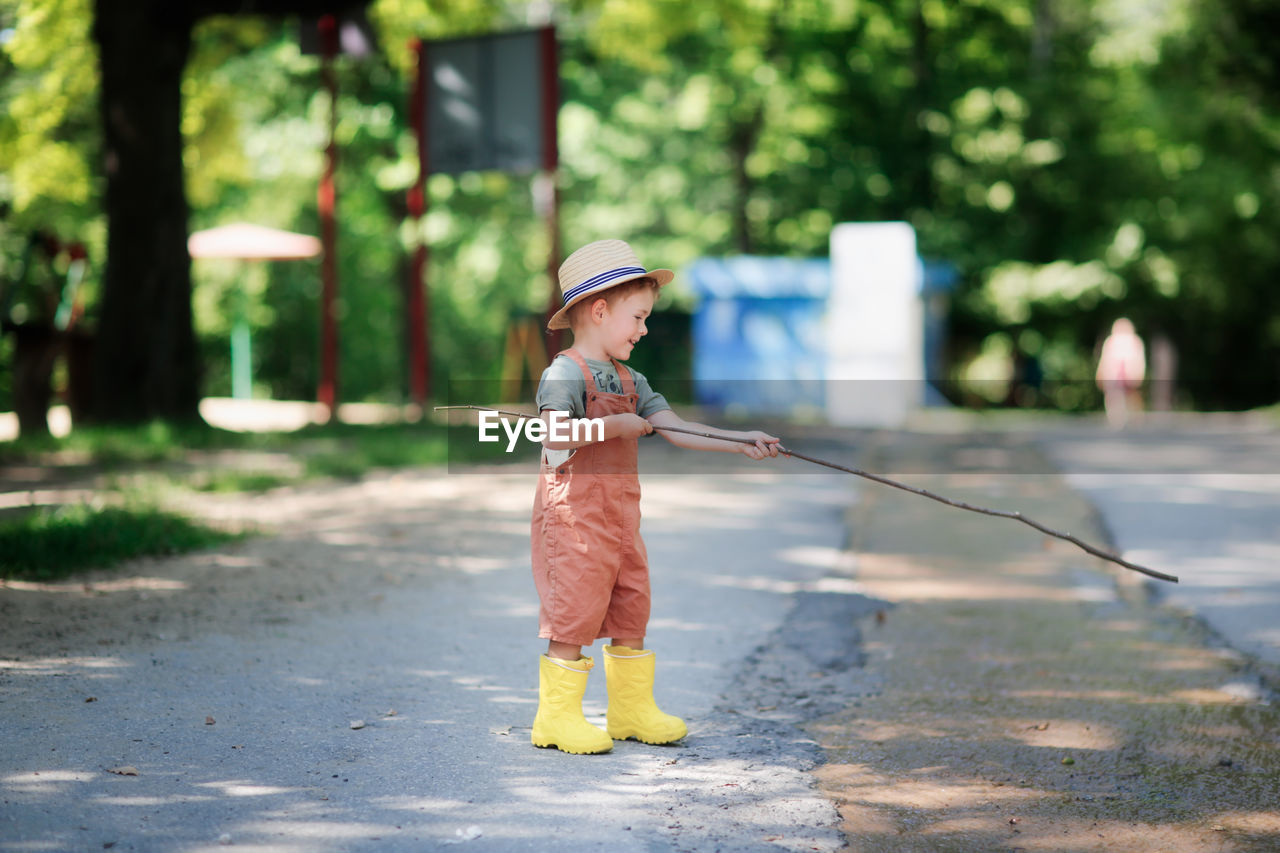 Cute european boy in hat and overalls in summer with stick and fishing rod plays with stream on road 